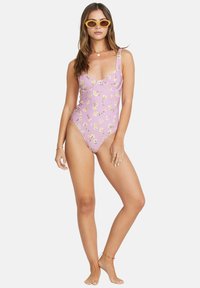 Maillot de bain une pièce rose avec un motif floral jaune, présentant un décolleté en V, des bretelles larges et un design ajusté. Accompagné de lunettes de soleil jaunes.