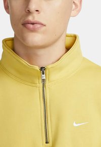 Fleece giallo con zip alta, caratterizzato da una zip argentata e un piccolo logo Nike bianco sul petto. Texture liscia.