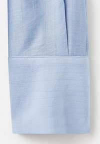 Manche de chemise bleu clair avec un large poignet présentant une texture fine tissée et des coutures visibles le long du bord du poignet.