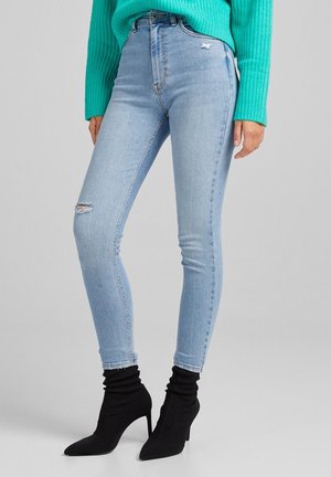 Jean skinny bleu clair avec détailles déchirées aux genoux, taille haute et coupe ajustée. Associé avec des bottines noires à talons.