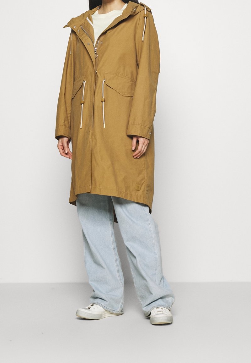 Długi trencz w kolorze khaki z kapturem na sznurku, dwoma przednimi kieszeniami i regulowanymi troczkami. Połączony z jasnoniebieskimi szerokimi jeansami i białymi trampkami.