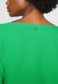 Zelený žebrovaný top s širokým výstřihem. Významný detail: malý logový akcent na zádech, texturovaná látka a živá barva.