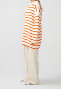 Orange-weiß gestreifter Pullover mit Knöpfen an der Schulter, kombiniert mit lockeren cremefarbenen Hosen und weißen Turnschuhen. Glatte Textur.