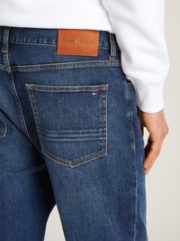Jeans de ganga azul escuro com um rótulo de pele TOMMY HILFIGER na cintura nas costas. As características incluem dois bolsos traseiros e um corte reto.