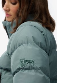 Pufferjakke i afdæmpet teal med en skinnende finish. Har en høj krave, polstrede ærmer og broderet logo i mørkegrøn på ærmet.