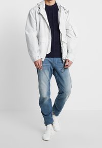 Vit lättviktig jacka med texturerade accenter, tillsammans med en marinblå stickad tröja, blå slitna jeans och vita sneakers.