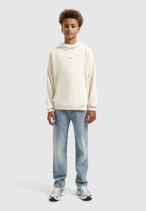 Teenager trägt einen cremefarbenen Hoodie mit kleinem blauem Text, hellen Jeans und weißen Sneakers, steht vor einem schlichten Hintergrund.