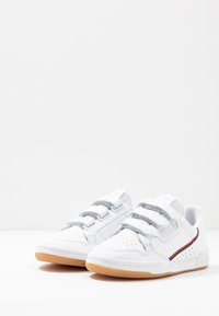 Vita sportskor med en slät läderöverdel, grå kardborreband, perforeringar och en sula av gummigummi. Röd och grön accentrand.