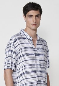 Košela s krátkym rukávom, so zapínaním na gombíky z bielej látky, ktorá má horizontálne pruhy v námorníckom modrom. Výrazné detaily zahŕňajú golier a uvoľnený strih.