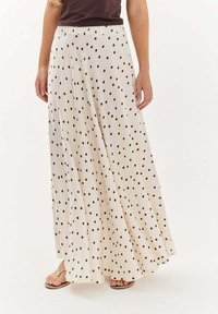 Žena v priesvitnej bielej maxi sukni s čiernymi bodkami, skombinovanej s tmavým topom a béžovými sandálmi.