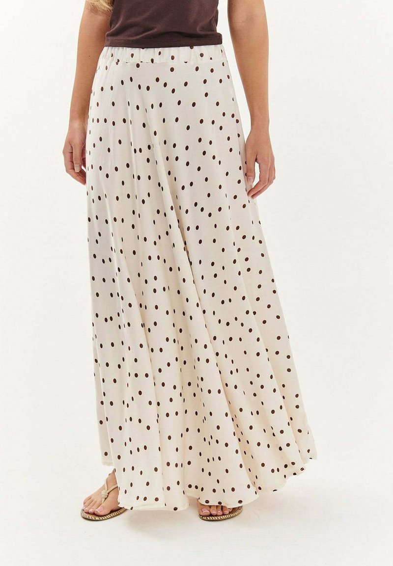 Žena v priesvitnej bielej maxi sukni s čiernymi bodkami, skombinovanej s tmavým topom a béžovými sandálmi.
