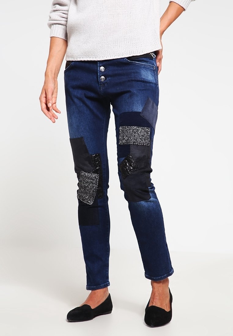 Jeans de mezclilla azul con varios parches de tela negra, incluyendo materiales texturizados y lisos, con un diseño ajustado y dos botones en la cintura.