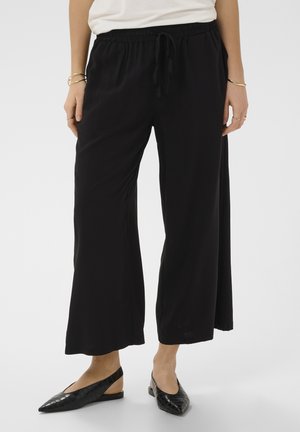 Kobieta ubrana w luźne, czarne spodnie z szerokimi nogawkami i elastyczną talią oraz czarne, spiczaste buty na niskim obcasie typu slingback na jednolitym tle.