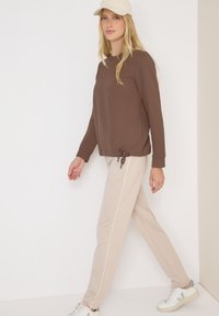 Felpa marrone a trama con coulisse laterale, abbinata a pantaloni beige con strisce bianche ai lati e sneakers bianche. Cappellino beige.