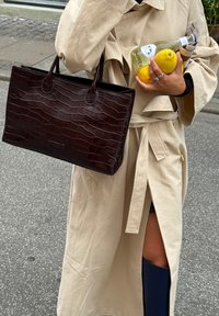 Borsa tote in pelle marrone scuro con motivo coccodrillo, con due manici, portata insieme a un limone giallo e una bottiglia, indossata con un trench beige.