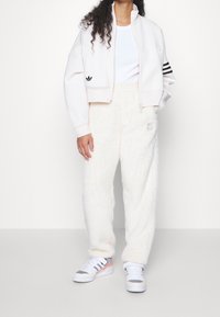 Lyhennetty valkoinen fleece-takki, jossa musta adidas-logo ja kolme mustaa raitaa hihoissa, yhdessä kermanväristen pörröisten housujen ja valkoisten lenkkareiden kanssa.