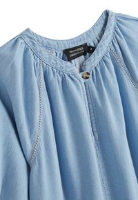 Chemise bleu clair en denim doux avec un col rond, fronces aux épaules et un bouton décoratif à l'avant. Étiquette visible.