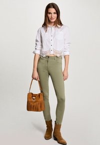 Weißes, kurzes Hemd mit Spitzenabschluss, olivgrüne Skinny-Jeans, braune Wildleder-Stiefeletten und eine braune Fransen-Tasche.