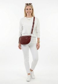 Naine valges riietuses ja valgete tennistega, kandmas pruuni nahast ristihoidjat koos päikeseprillidega peas, naeratades.