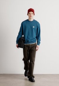 Felpa blu con un piccolo logo, pantaloni verde oliva e scarpe nere. Il modello indossa un berretto rosso e tiene in mano una giacca nera.