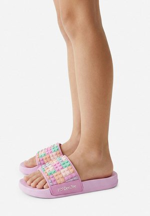 Feet wearing pastel-colored slide sandals with raised building block patterns and "I heart DeeZee" text on the straps.