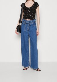 Černý kratší top s květinovými vzory a volánkovými rukávy, kombinovaný s vysoko pasovými, širokými modrými džíny a černými sandály na řemínky.