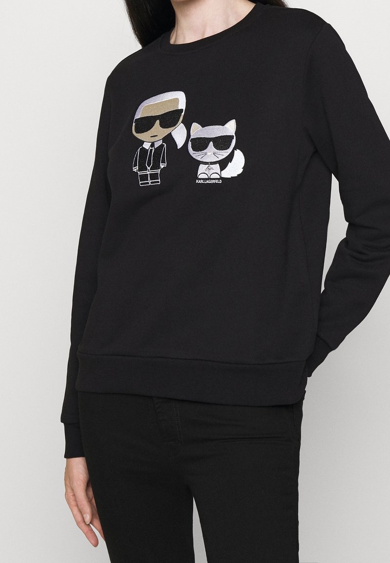 Sweat-shirt noir avec des personnages de dessin animé d'un homme et d'un chat portant des lunettes de soleil, centrés sur la poitrine.