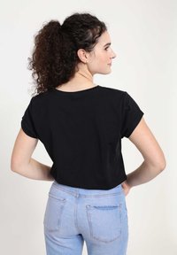 Žena s kudrnatými vlasy svázanými vzadu, oblečená v černém tričku s krátkým rukávem a světle modrých džínách, stojí s rukama na bocích, otočená zády.