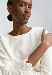 Maglione bianco texturizzato con scollatura ampia e polsini a boucle, caratterizzato da accenti arricciati sulle maniche. Tessuto liscio.
