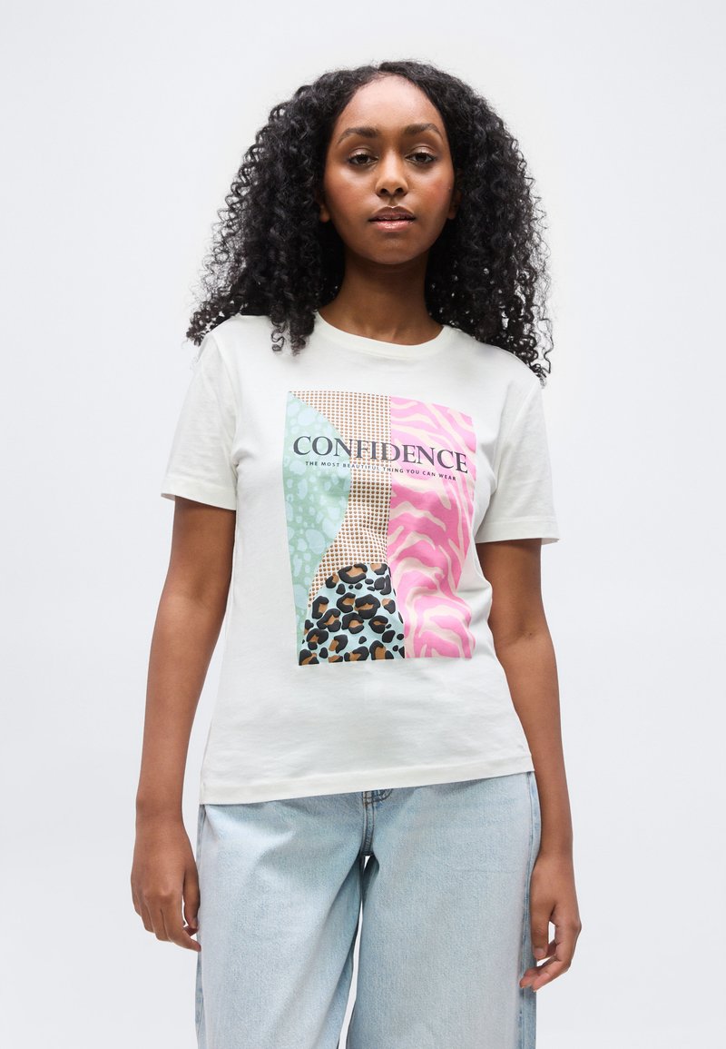 Γυναίκα με σγουρά μαλλιά που φοράει λευκό μπλουζάκι με πολύχρωμες ζωγραφιές ζώων και τη λέξη «ΑΥΤΟΠΕΠΟΙΘΗΣΗ», συνδυασμένο με ανοιχτό μπλε τζιν.