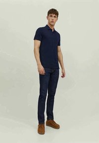 Camisa pólo azul-marinho de mangas curtas, combinada com jeans de ganga escura e sapatos de camurça castanhos. O modelo está numa fundo neutro.