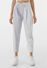 Světlé šedé kalhoty s kombinací kostkované a vertikálně pruhované textury, s boxovaným předním dílem, zkrácenou délkou a vysokým pasem. Spárované s bílými teniskami.