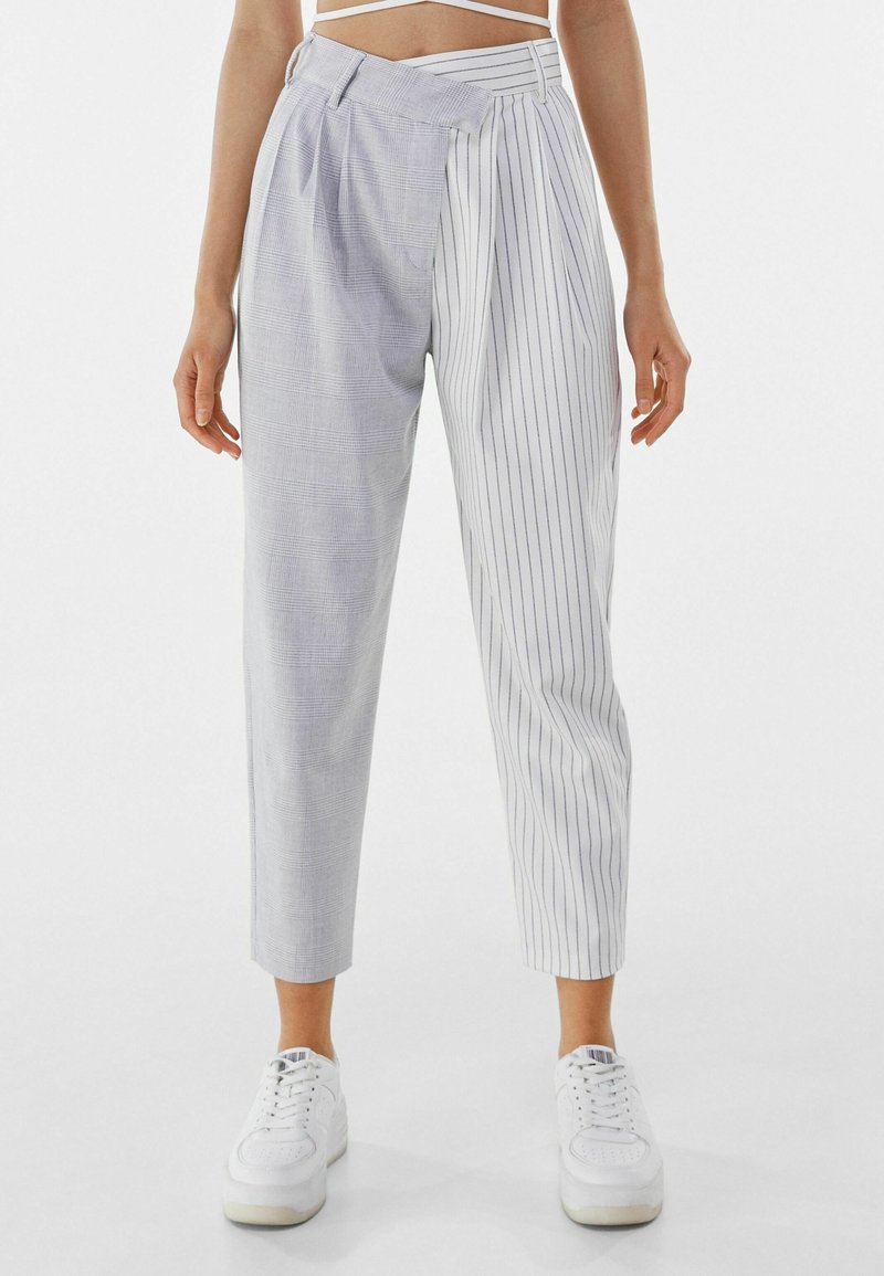 Světlé šedé kalhoty s kombinací kostkované a vertikálně pruhované textury, s boxovaným předním dílem, zkrácenou délkou a vysokým pasem. Spárované s bílými teniskami.