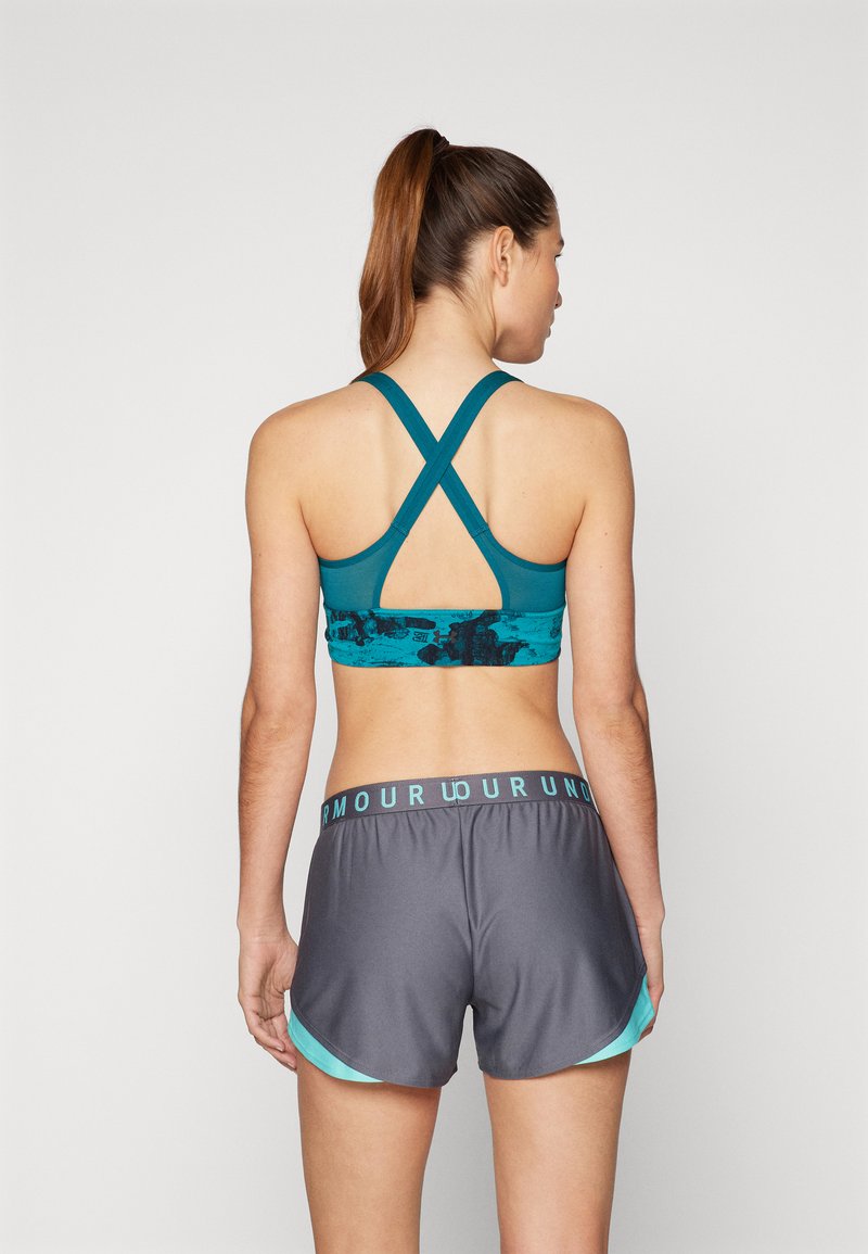 Reggiseno sportivo teal con design a incrocio sulla schiena e motivo astratto, abbinato a pantaloni sportivi grigi con vita teal e dettagli laterali.