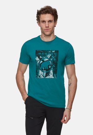 Man in een teal Mammut t-shirt met mammoetlogo en zwarte broek, staande met één hand in de zak op een lichte achtergrond.