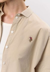 Beige skjorta med knappar och mjuk textur, med ett broderat motiv av en ryttare på vänstra bröstet samt knappdetaljer på knappslån.