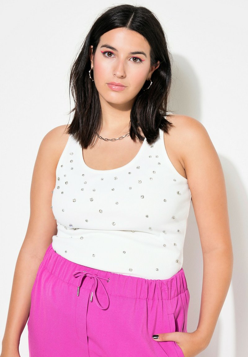 Camiseta sin mangas de rayas blancas con tachuelas plateadas, combinada con pantalones de chándal rosas con cordón. La parte superior tiene un escote redondeado y una silueta ajustada.