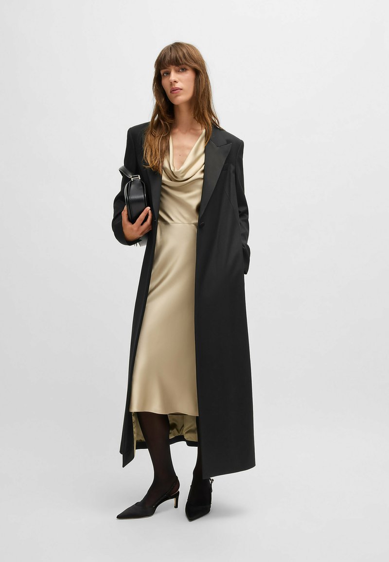 Un long manteau noir sur une robe en soie dorée douce avec un col drapé. Le mannequin tient un petit sac à main noir et porte des talons aiguilles noirs ainsi que des collants.