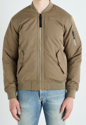 Personne portant une veste bomber marron zippée avec poignets et ourlet côtelés, deux poches avant à rabat et une poche zippée sur la manche gauche.