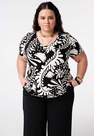 Vrouw met lang, donker haar, gekleed in zwarte broek en een zwart-witte, gebloemde korte mouw top, staand met handen in de zakken.