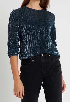 Mujer con un top de lentejuelas azul oscuro de manga larga y vaqueros negros de talle alto con una franja roja en el lateral.