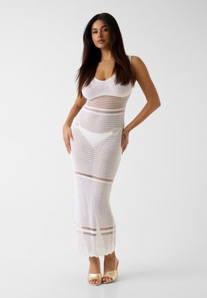 Kobieta z długimi ciemnymi włosami, ubrana w białą sukienkę na szydełku bez rękawów na białą bieliznę, nosząca złote sandały na odkryte palce, stoi z rękami na biodrach.