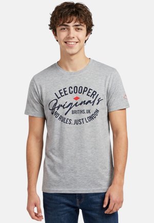Jeune homme souriant, portant un t-shirt gris clair avec le texte "LEE COOPER Originals BRITISH.UK NO RULES, JUST LONDON", jean bleu, sur fond uni.