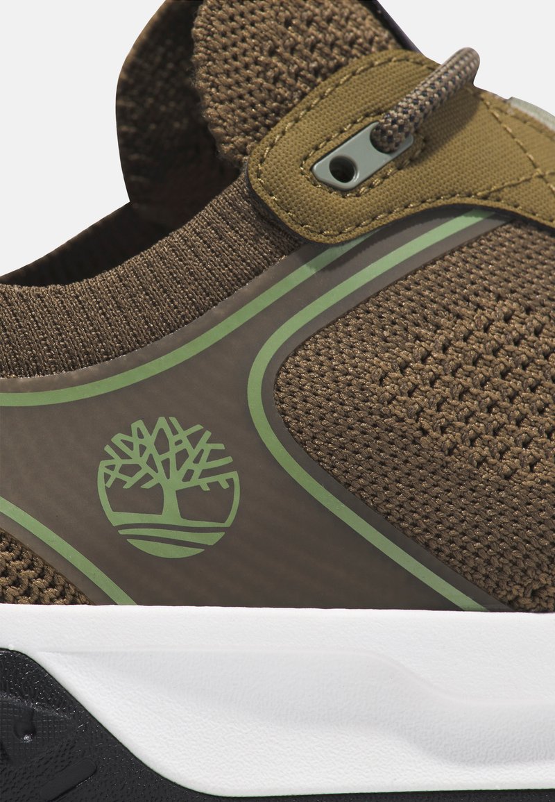 Zapatilla marrón con parte superior de punto texturizado, acentos verdes y suela blanca. Presenta un logo en el centro y un lazo para facilitar su uso.