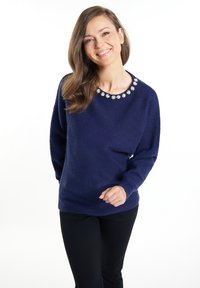 Marineblauem Pullover mit langen Ärmeln, strukturiertem Stoff, floralen Verzierungen am Ausschnitt, kombiniert mit schwarzen Hosen.