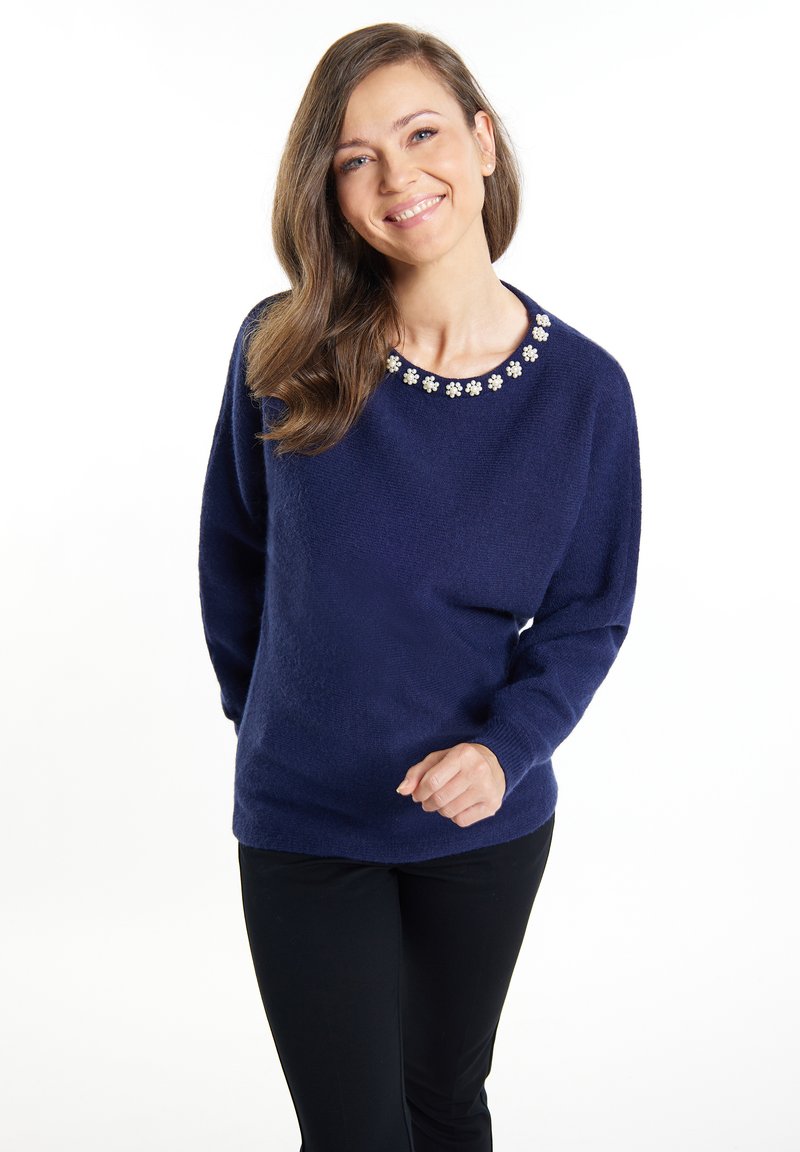 Marineblauem Pullover mit langen Ärmeln, strukturiertem Stoff, floralen Verzierungen am Ausschnitt, kombiniert mit schwarzen Hosen.