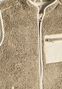 Gilet in pile beige con una superficie texturizzata e pelosa. Presenta una chiusura con zip e una piccola tasca con zip impreziosita da un accento beige.