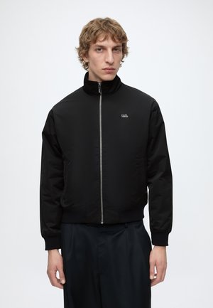 Jeune homme aux cheveux bouclés portant une veste noire zippée et un pantalon foncé, debout devant un fond clair uni.
