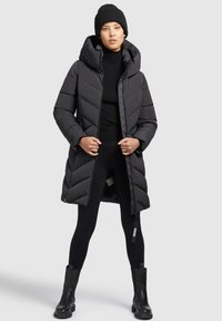 Doudoune noire avec capuche, présentant un design matelassé et une fermeture éclair, associée à des leggings noirs et des bottines.