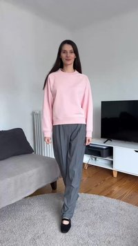 Woman with long dark hair wearing a light pink sweatshirt, gray pants, and black shoes standing on a gray rug in a living room.